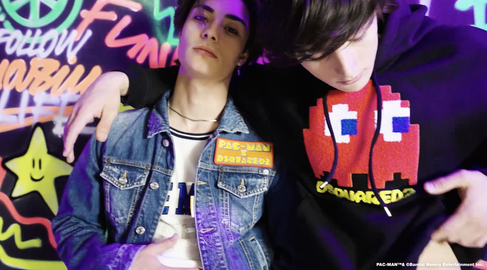 Two teenagers hugging on a graffiti wall background. One wears a leather jacket and the other one wears a black hoodie with pac-man characters on it.