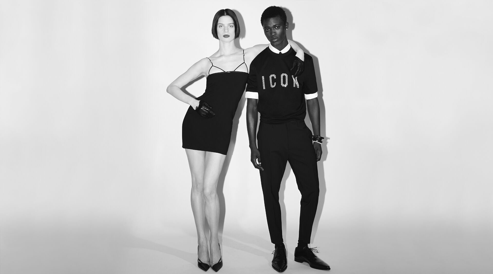 picture in black and white of man and woman. The man is wearing a black tshirt with white ICON logo and black elegant pants. the woman is wearing a black tight dress with high heels. 
