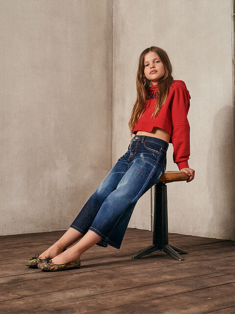 girl with long brown hair, red long sleeve shirt and wide denim jeans sitting on a chair
