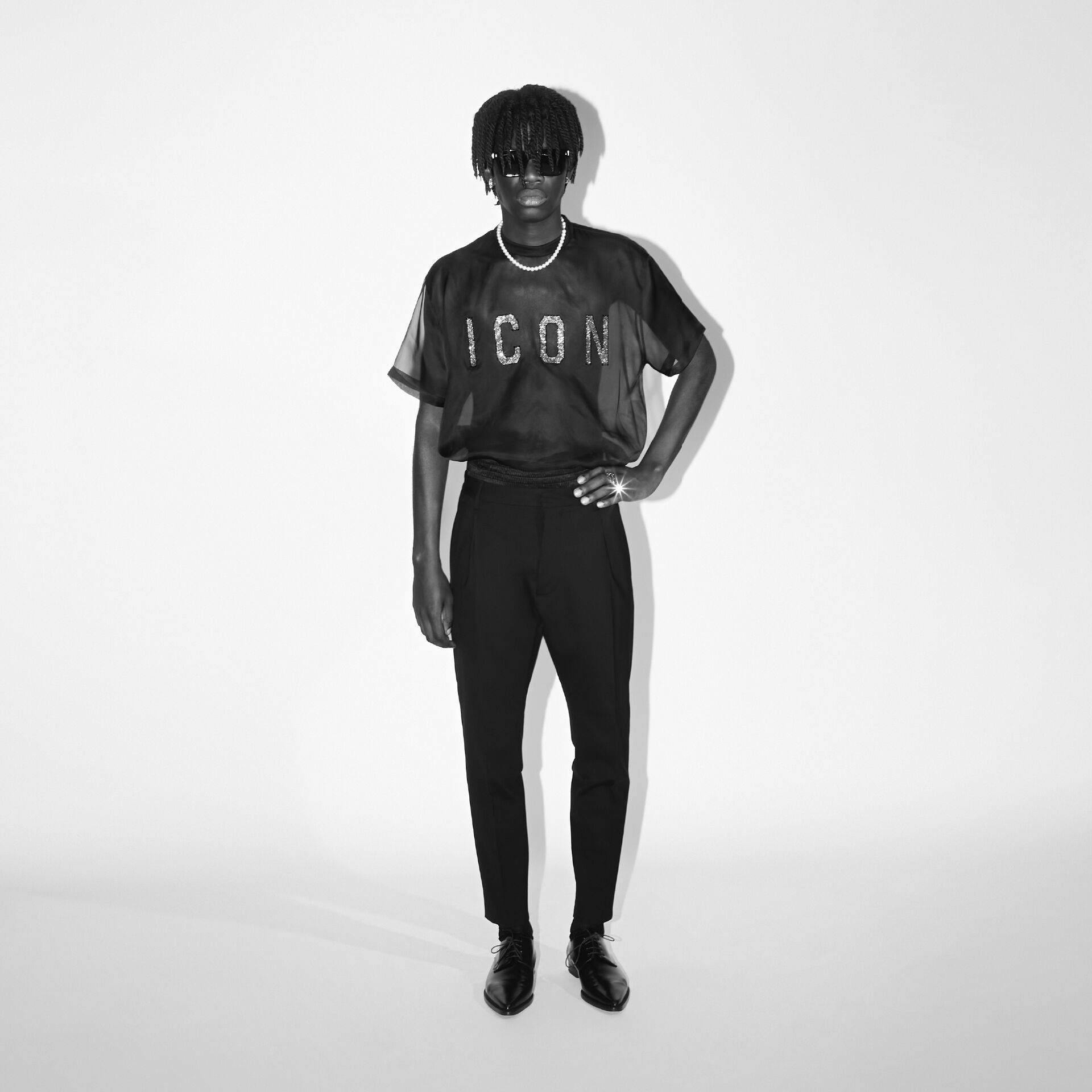 Picture in black and white of a man wearing a transparent black t-shirt with ICON logo on it and black classic pants