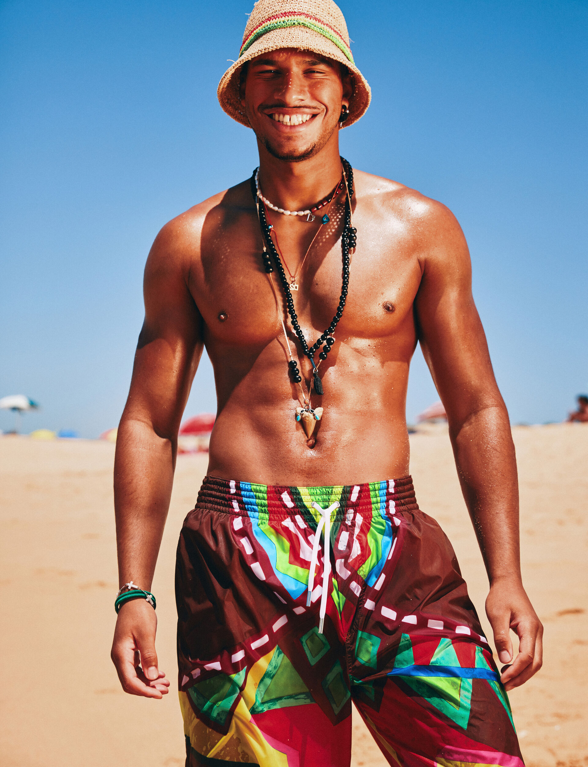 Men at the beach wearing a light brown hat and colorful swim trunks from dsquared2 ss23 beachwear collection