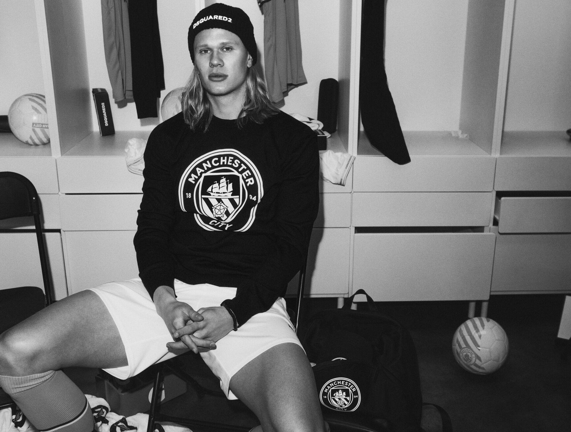 picture in black and white of erling haaland sitting on a chair in the locker room wearing a black sweater with man city logo from dsquared2 x manchester city capsule collection