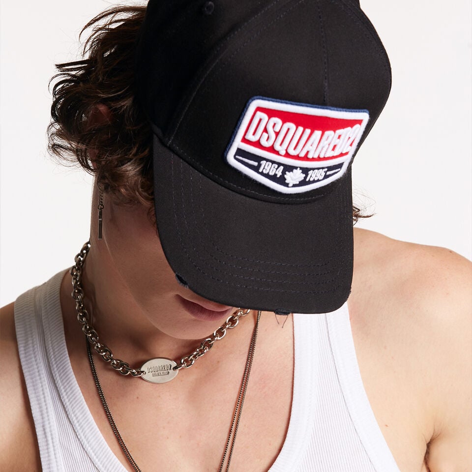 Man with black baseball cap with dsquared2 patch logo on it, wears a white tank top and long necklaces. 