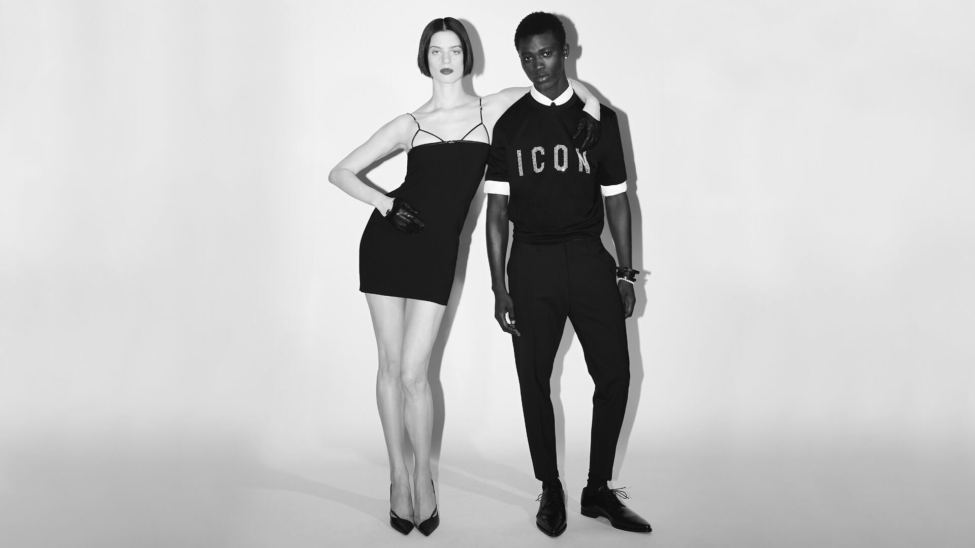 picture in black and white of man and woman. The man is wearing a black tshirt with white ICON logo and black elegant pants. the woman is wearing a black tight dress with high heels. 
