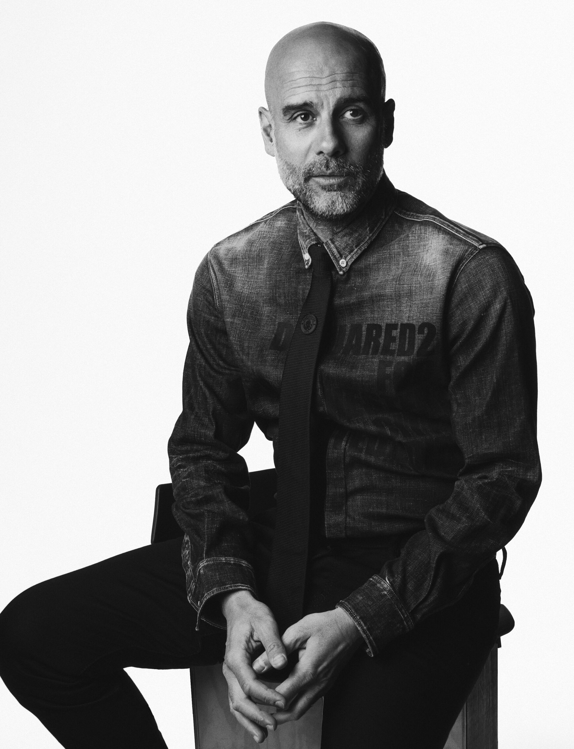 picture in black and white of pep guardiola sitting on a chair and wearing a denim shirt from dsquared2 x manchester city capsule collection